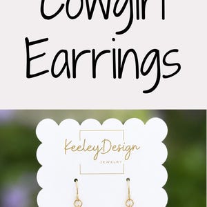 May include: Three pairs of dangling earrings shaped like cowboy boots on white scalloped display cards labeled Keeley Design Jewelry. The boots feature black, pink with white floral patterns, and grey with white star patterns, all with tan soles.