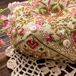 Pode incluir: Uma almofada quadrada bege com intrincados bordados florais em ponto cruz em tons de vermelho, verde e rosa. A almofada é adornada com pequenos botões redondos rosa claro e contas. Descansa sobre uma toalha de mesa bege de crochê, com flores rosa em primeiro plano.