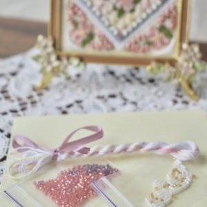 May include: A craft kit with a light yellow background. The kit includes a pink and white twisted cord, a pink ribbon, small bags of pink and purple beads, and a white beaded design with gold accents.