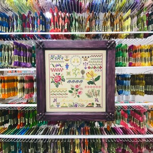 Pode incluir: Um padr&atilde;o de ponto cruz emoldurado com uma moldura roxa e um fundo branco. O padr&atilde;o apresenta uma variedade de desenhos florais e geom&eacute;tricos, incluindo uma coroa de flores, uma borboleta e um p&aacute;ssaro. O texto "Marie Berger" est&aacute; impresso na parte inferior do padr&atilde;o.