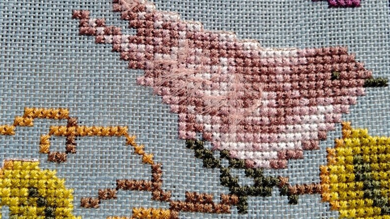 May include: Close-up of a cross-stitch embroidery featuring a pink flower with green leaves and a yellow flower with brown stems.
