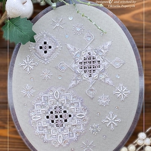 May include: Oval embroidery hoop with a detailed snowflake design. The design includes the text "Snowflakes" and "Designed and stitched by Viktoriia Popovych". The embroidery is in white thread on a light green fabric, with a wooden frame.