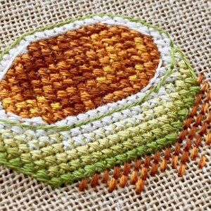 Puede incluir: Un primer plano de un dise&ntilde;o de bordado cosido a mano sobre tela de arpillera. El dise&ntilde;o presenta una forma circular con anillos conc&eacute;ntricos de puntadas de color naranja, blanco y verde.