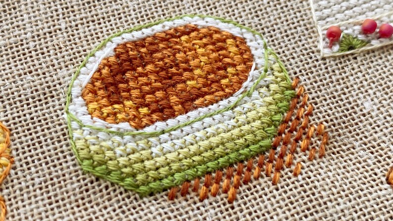 Puede incluir: Un primer plano de un dise&ntilde;o de bordado cosido a mano sobre tela de arpillera. El dise&ntilde;o presenta una forma circular con anillos conc&eacute;ntricos de puntadas de color naranja, blanco y verde.