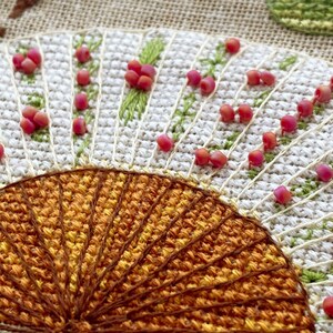 Puede incluir: Primer plano de un dise&ntilde;o de bordado cosido a mano que presenta un motivo en forma de abanico con hilos naranja y marr&oacute;n. El abanico est&aacute; rodeado de un fondo blanco con cuentas rosadas y puntadas verdes.