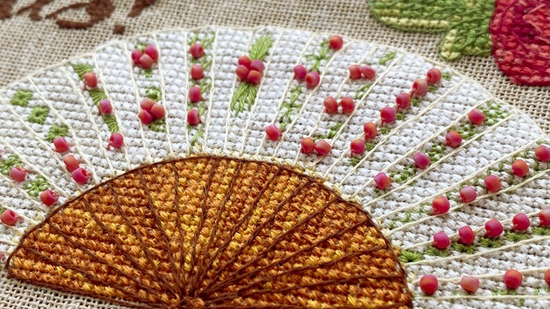 Puede incluir: Primer plano de un dise&ntilde;o de bordado cosido a mano que presenta un motivo en forma de abanico con hilos naranja y marr&oacute;n. El abanico est&aacute; rodeado de un fondo blanco con cuentas rosadas y puntadas verdes.