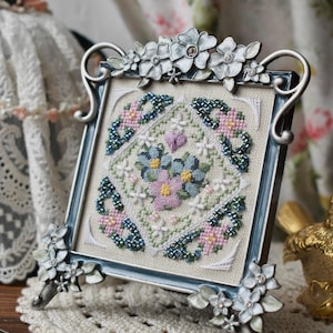 Pode incluir: Um design floral bordado em ponto cruz enquadrado. A obra de arte quadrada apresenta um grupo central de flores azuis, rosas e amarelas, rodeado por um padrão geométrico em azul e rosa. A moldura é azul clara com detalhes florais prateados.