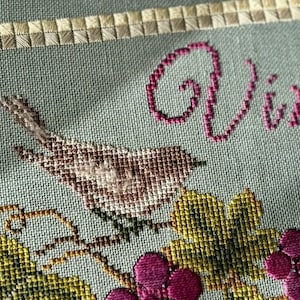 May include: Cross-stitch embroidery of a bird perched on a vine with grapes. The word "Vive" is stitched in pink above the bird.