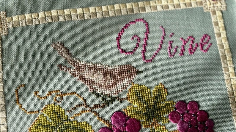 May include: Cross-stitch embroidery of a bird perched on a vine with grapes. The word "Vive" is stitched in pink above the bird.