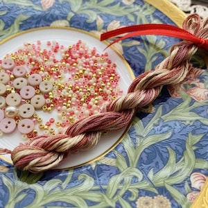 May include: A close-up of a white ceramic plate with a floral design. The plate is filled with pink and white buttons, gold and pink beads, and a long, braided strand of red and cream colored embroidery floss.