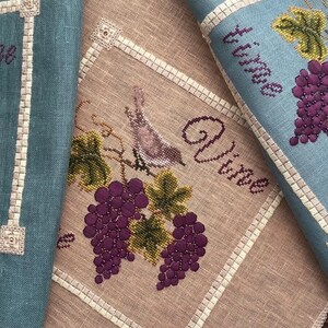 May include: Two embroidered kitchen towels with a blue and beige background. Each towel features a bird perched on a vine with grapes. The text "Vine" is embroidered on each towel.