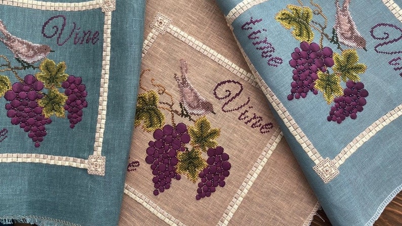 May include: Two embroidered kitchen towels with a blue and beige background. Each towel features a bird perched on a vine with grapes. The text "Vine" is embroidered on each towel.