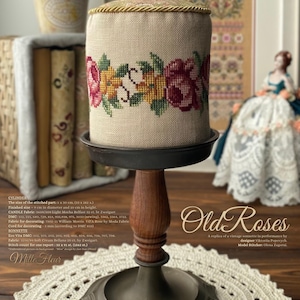 Peut inclure: Un cylindre en point de croix beige avec un motif floral de roses jaunes et roses, brodé sur un tissu de couleur crème. Le cylindre est présenté sur un support en bois avec une base noire. Le texte "Old Roses" est écrit dans une police décorative sous le cylindre.