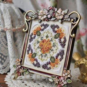 Pode incluir: Uma peça decorativa bordada em ponto cruz, emoldurada, com um design floral em tons de laranja, verde e roxo. A moldura castanha escura tem detalhes dourados e enfeites florais. Exibida sobre uma toalha de renda creme.