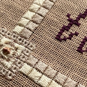 May include: Close-up of a beige burlap fabric with white and brown embroidery. The embroidery features a square design with a small, shiny, copper-coloured bead in the centre.