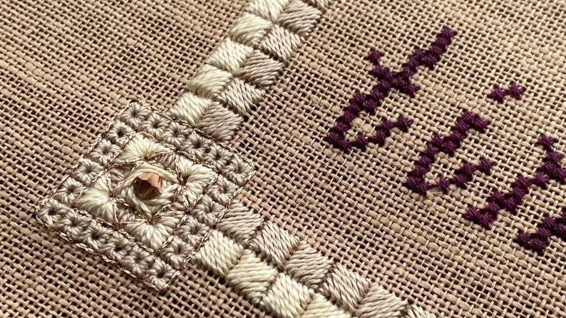 May include: Close-up of a beige burlap fabric with white and brown embroidery. The embroidery features a square design with a small, shiny, copper-coloured bead in the centre.