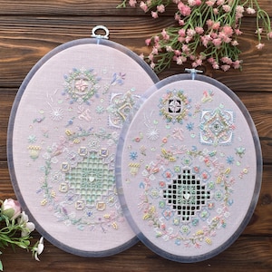 Könnte beinhalten: Zwei ovale Stickrahmen mit aufwendigen floralen und geometrischen Designs. Die Stickerei zeigt Pastellfarben wie Rosa, Blau und Gelb auf einem hellrosa Stoff. Die Rahmen werden auf einer Holzoberfläche präsentiert.