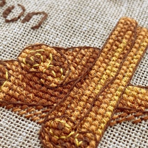 Puede incluir: Primer plano de un dise&ntilde;o de bordado de punto de cruz que presenta una imagen estilizada de palitos de canela en tonos marrones y amarillos.