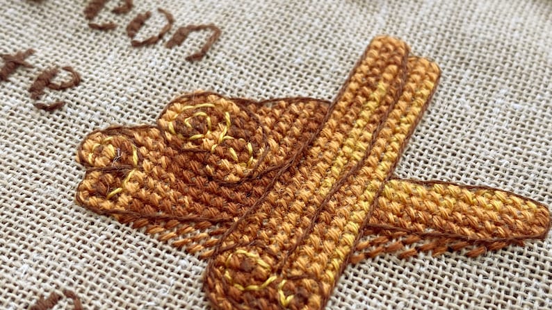 Puede incluir: Primer plano de un dise&ntilde;o de bordado de punto de cruz que presenta una imagen estilizada de palitos de canela en tonos marrones y amarillos.