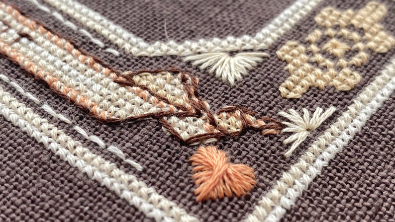 Pode incluir: Close-up de um tecido castanho com bordados brancos e castanhos. O bordado apresenta uma variedade de formas, incluindo cora&ccedil;&otilde;es, estrelas e flores.