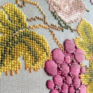 May include: Close-up of a cross-stitch embroidery featuring a vine with green leaves and pink grapes. The embroidery is on a white linen fabric.