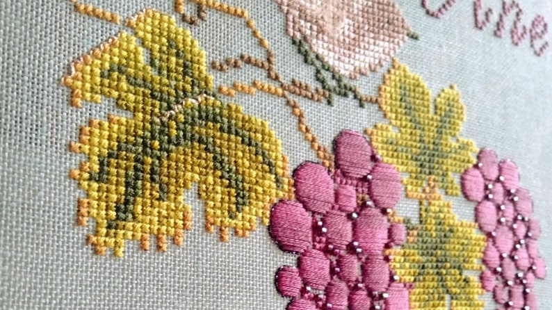 May include: Close-up of a cross-stitch embroidery featuring a vine with green leaves and pink grapes. The embroidery is on a white linen fabric.