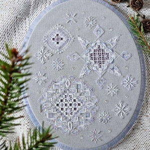 May include: An oval, light grey embroidered piece with detailed white snowflake and geometric patterns. The embroidery is framed in light blue, giving a wintery, handcrafted feel. Small, sparkling embellishments add to the design. The piece is a decorative item.