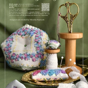 Pode incluir: Uma exibição de artigos de bordado em ponto cruz, incluindo um alfineteiro, um porta-agulhas e tesouras. O design apresenta a palavra "Hydrangea" e um padrão floral em azul, rosa e roxo. O tamanho do design é de 30 x 19 cm.