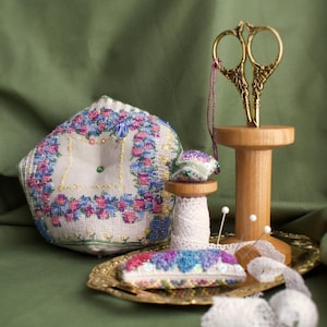 Pode incluir: Um kit de costura vintage com um almofadinha para alfinetes bordada com flores, um carretel de fio branco, um par de tesouras douradas e uma bandeja pequena com um almofadinha para alfinetes bordada com flores.