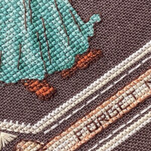 Pode incluir: Close-up de um bordado de ponto cruz com fundo castanho. O bordado apresenta uma figura verde e azul com botas castanhas e o texto "Forget Me Not" em fio branco e castanho.