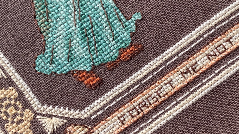 Pode incluir: Close-up de um bordado de ponto cruz com fundo castanho. O bordado apresenta uma figura verde e azul com botas castanhas e o texto "Forget Me Not" em fio branco e castanho.