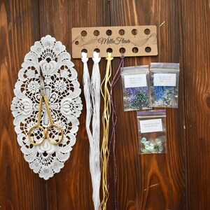 May include: A Mille Fleur embroidery kit with white, yellow, and purple thread, three bags of beads, a pair of gold scissors, and a white lace doily.