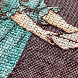 Pode incluir: Close-up de um bordado de ponto cruz de uma pessoa vestindo um vestido turquesa e sapatos brancos. O bordado est&aacute; em um tecido marrom.