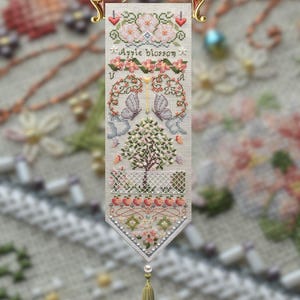 May include: A cross-stitch banner with the words "Apple Blossom" and the numbers "20" and "25". The banner features floral and butterfly motifs, a tree, and a decorative tassel. The banner is held by a gold-coloured metal hanger.