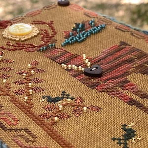 May include: Close-up of a detailed, hand-stitched textile art piece. The piece features a tan background with intricate embroidery in shades of brown, red, and green. Decorative elements include a heart-shaped button, beaded accents, and a decorative button.