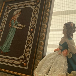 Pode incluir: Um bordado emoldurado com uma menina segurando uma carta e o texto "St. Valentine's Day Forget Me Not". Uma boneca de porcelana com um vestido de renda branca e flores cor-de-rosa est&aacute; em primeiro plano.