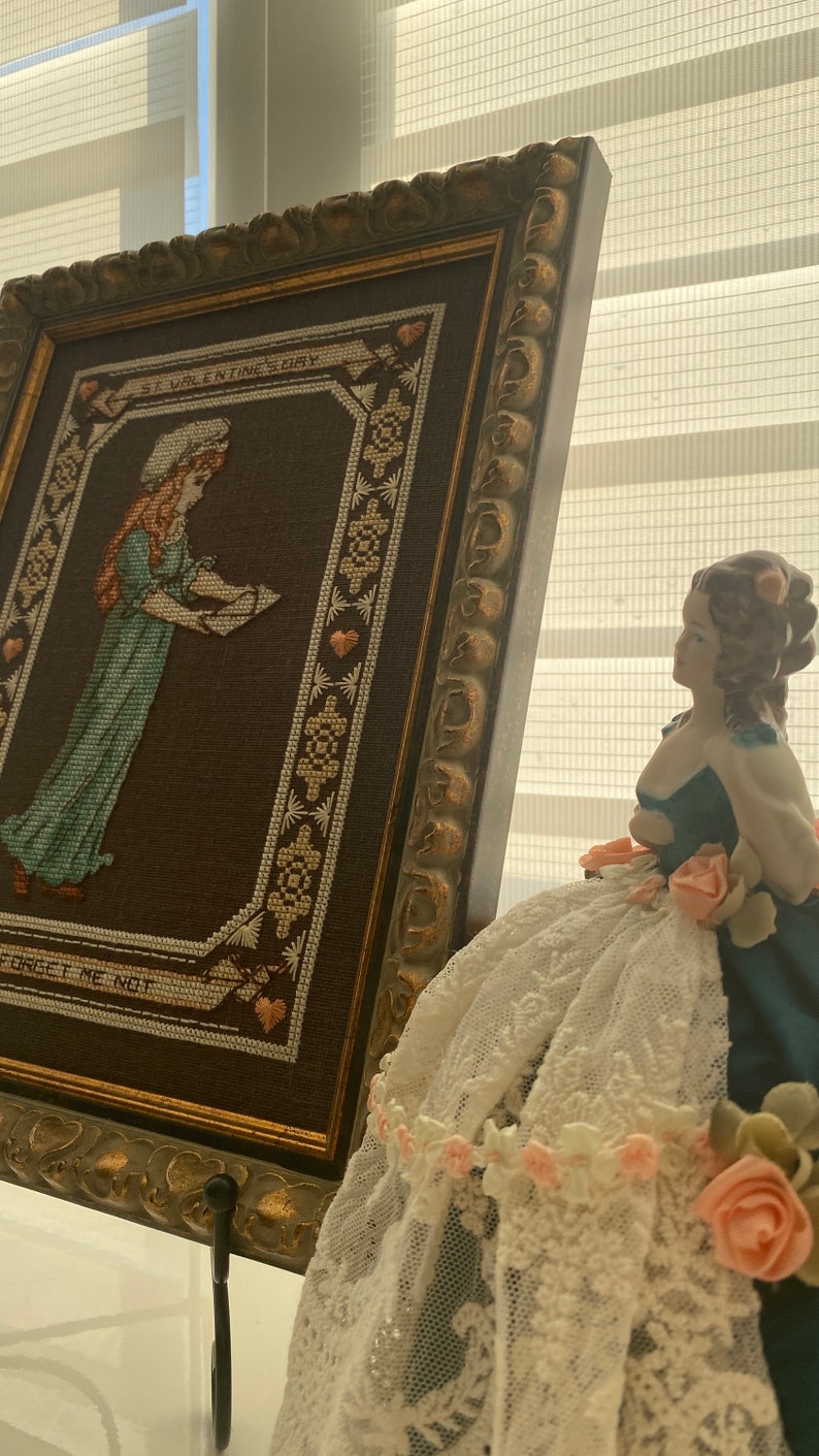 Pode incluir: Um bordado emoldurado com uma menina segurando uma carta e o texto "St. Valentine's Day Forget Me Not". Uma boneca de porcelana com um vestido de renda branca e flores cor-de-rosa est&aacute; em primeiro plano.