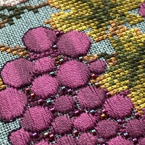 May include: Close-up of a cross-stitch embroidery design featuring a cluster of purple grapes with small beads sewn around them. The background is a light blue fabric with a yellow and green cross-stitch pattern.