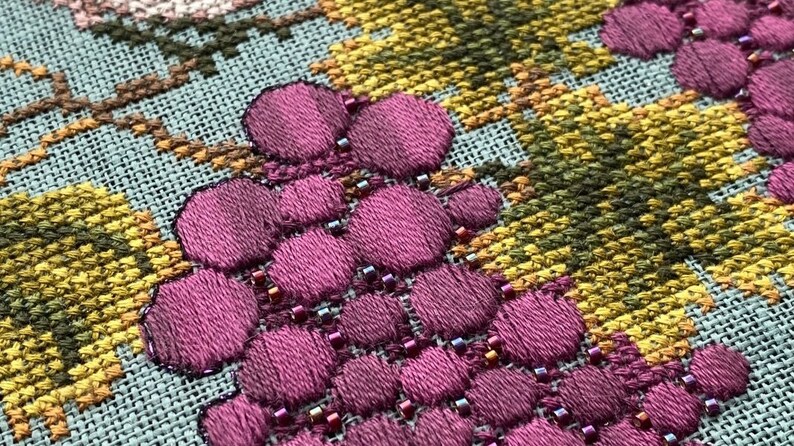 May include: Close-up of a cross-stitch embroidery design featuring a cluster of purple grapes with small beads sewn around them. The background is a light blue fabric with a yellow and green cross-stitch pattern.