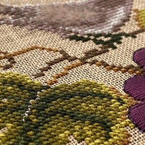 May include: Close-up of a cross-stitch embroidery featuring a floral design with green leaves, brown vines, and purple grapes with dark purple beads.
