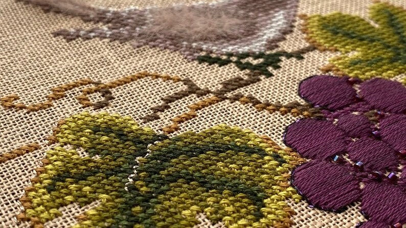 May include: Close-up of a cross-stitch embroidery featuring a floral design with green leaves, brown vines, and purple grapes with dark purple beads.