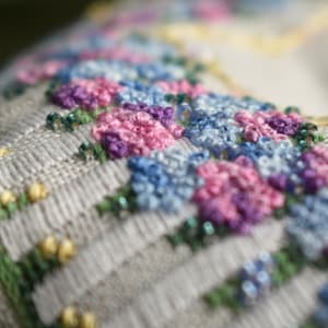 Pode incluir: Close-up de um tecido branco com flores bordadas em tons de rosa, azul e roxo. As flores estão dispostas em um padrão e têm um aspecto delicado, costurado à mão.