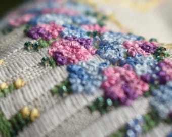 May include: Close-up of a white fabric with embroidered flowers in shades of pink, blue, and purple. The flowers are arranged in a pattern and have a delicate, hand-stitched look.