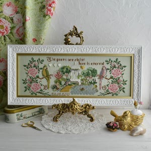 May include: A framed cross-stitch artwork with a white frame, featuring a garden scene with birds, roses, and the text "The poets are right: Love is eternal." The artwork is displayed on a white surface with decorative gold accents.