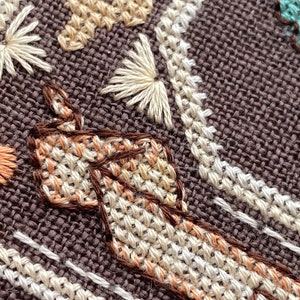 Pode incluir: Close-up de um tecido marrom com bordados coloridos. O bordado inclui um cora&ccedil;&atilde;o, uma flor e um desenho em forma de m&atilde;o.