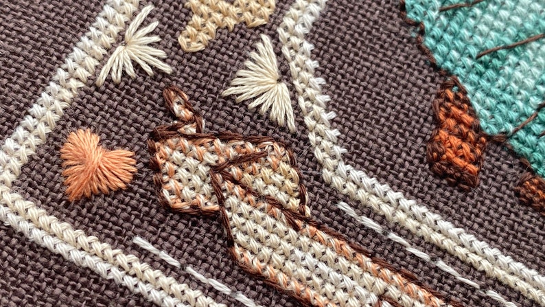 Pode incluir: Close-up de um tecido marrom com bordados coloridos. O bordado inclui um cora&ccedil;&atilde;o, uma flor e um desenho em forma de m&atilde;o.