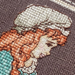 Pode incluir: Close-up de um bordado de ponto cruz que retrata uma mulher com cabelo castanho usando um chap&eacute;u branco e um vestido azul e verde.