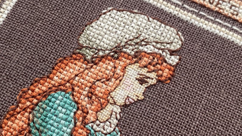 Pode incluir: Close-up de um bordado de ponto cruz que retrata uma mulher com cabelo castanho usando um chap&eacute;u branco e um vestido azul e verde.