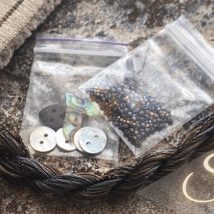 May include: A close-up of a craft kit with black and brown embroidery floss, a bag of iridescent beads, and a bag of buttons and shells. The word "Siren" is written in cursive on the bottom right corner of the image.