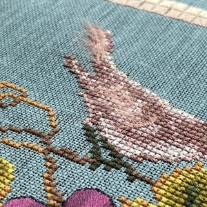 May include: Cross-stitch embroidery depicting a bird with pink and white feathers, perched on a vine with green leaves. The background is a light blue fabric.
